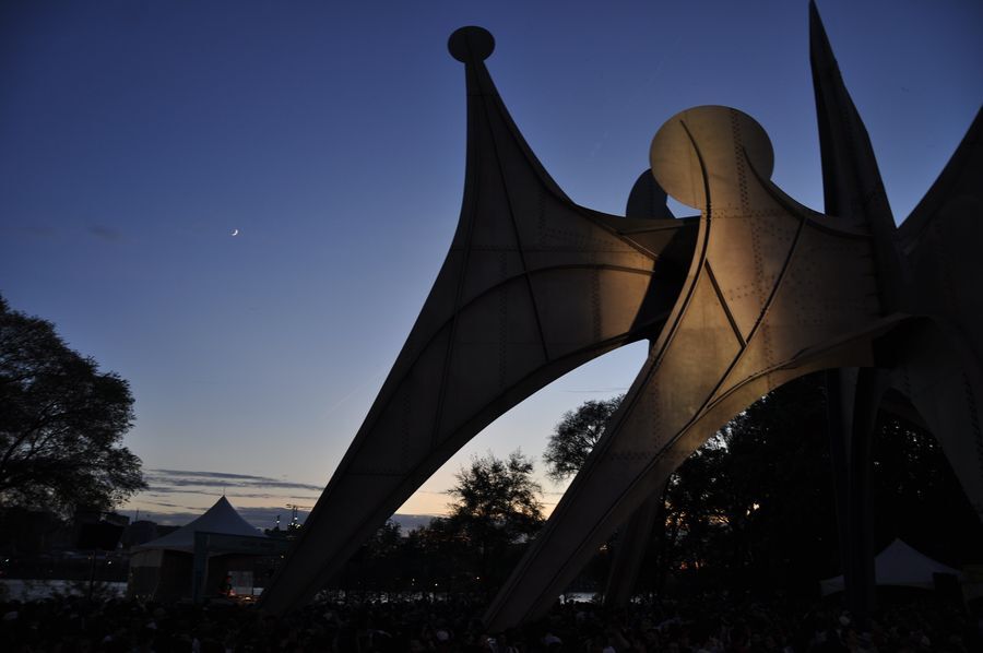 A massive metal sculpture appears in the evening dusk, lit by spotlights from the ground. A crescent moon is visible in the evening sky.