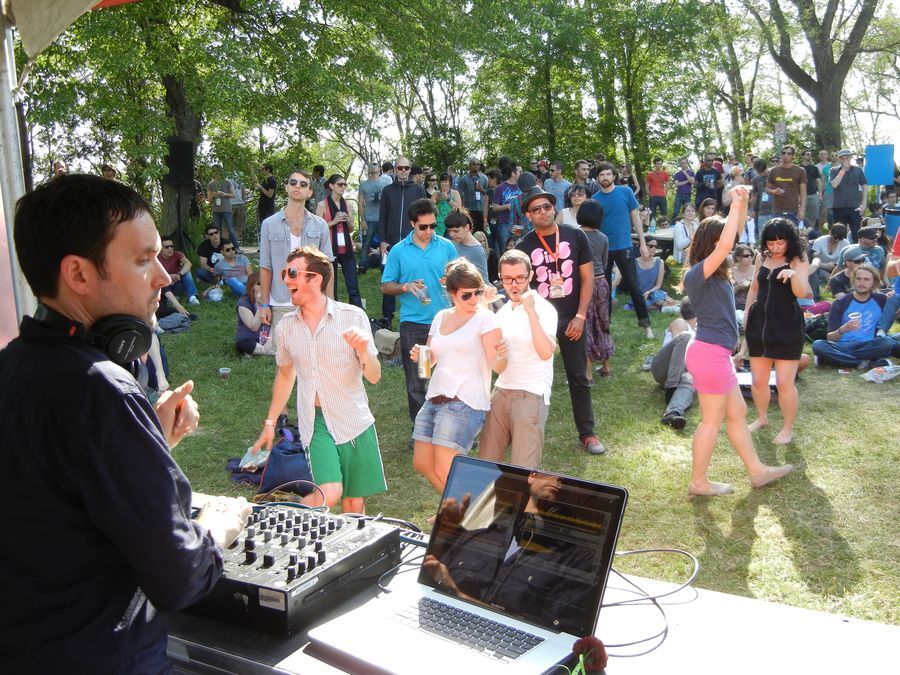 Sutekh, a short-haired man dressed in all black, touches knobs on a DJ mixer while glancing at a laptop. A small crowd of people dance with joy and abandon on a grassy field with trees behind them.